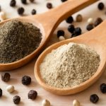 Black pepper powder and white pepper powder in wooden spoons on a rustic surface
