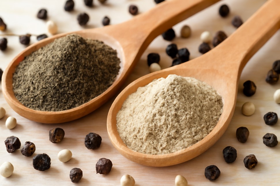 Black pepper powder and white pepper powder in wooden spoons on a rustic surface
