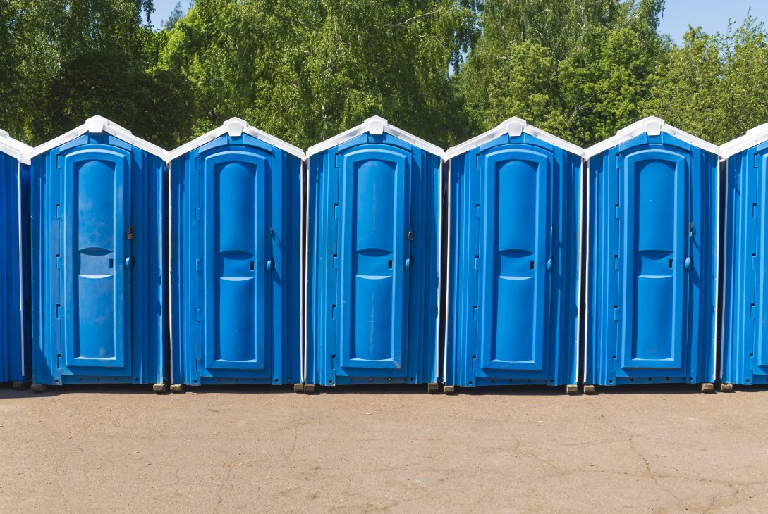 Row of blue portable toilets lined up outdoors at a construction site with green trees in the background