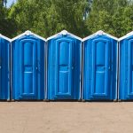 Row of blue portable toilets lined up outdoors at a construction site with green trees in the background