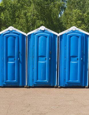 Row of blue portable toilets lined up outdoors at a construction site with green trees in the background
