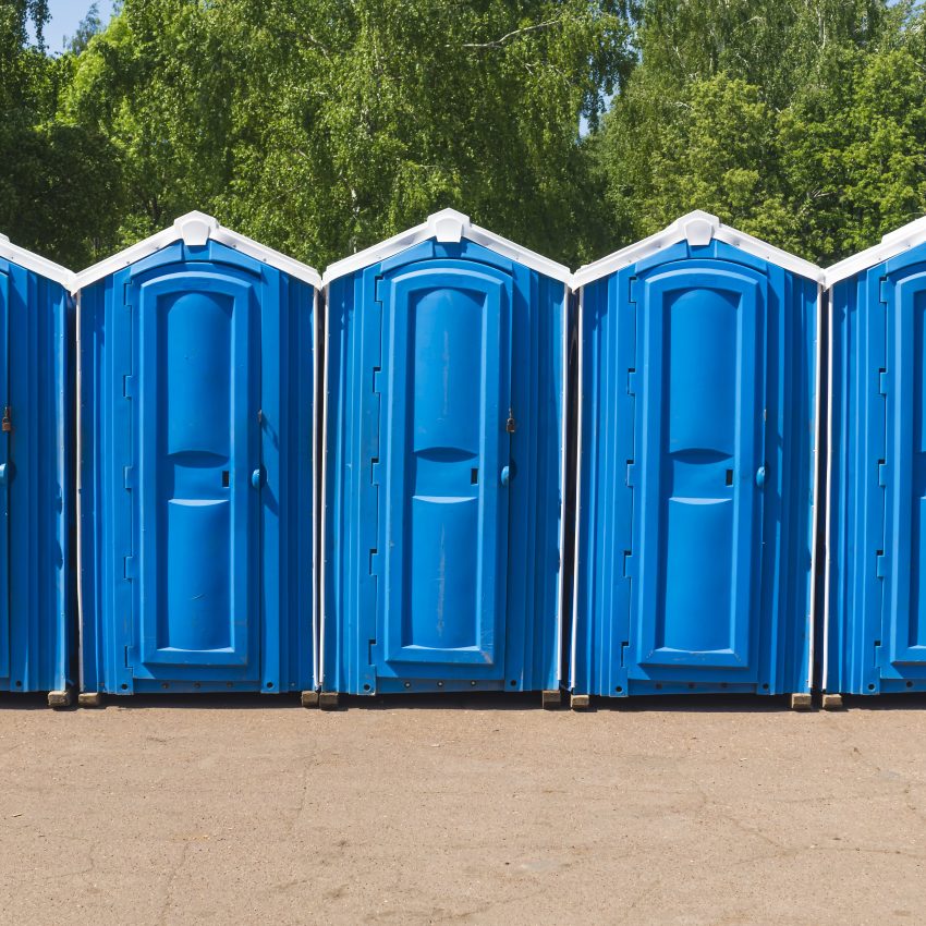 Row of blue portable toilets lined up outdoors at a construction site with green trees in the background