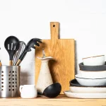Stylish dinner plates, cutlery set, and kitchen utensils on a wooden counter at white background.