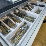 Organized kitchen drawer with stainless steel spoons, forks, and knives in a white cutlery tray.
