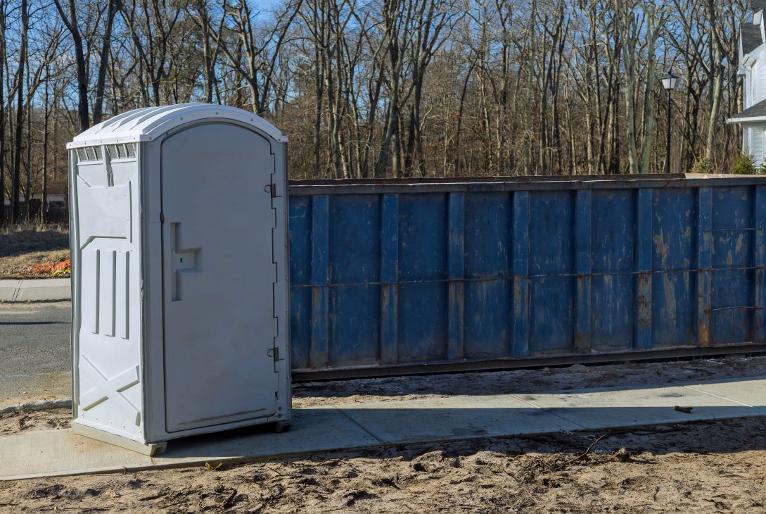 Single gray portable toilet placed at a residential construction site with a blue dumpster and bare trees in the background