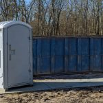 Single gray portable toilet placed at a residential construction site with a blue dumpster and bare trees in the background