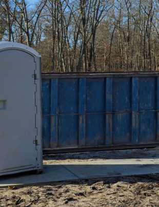 Single gray portable toilet placed at a residential construction site with a blue dumpster and bare trees in the background