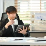 Young businessman working from home on a tablet, drinking coffee, embodying flexibility and financial freedom.